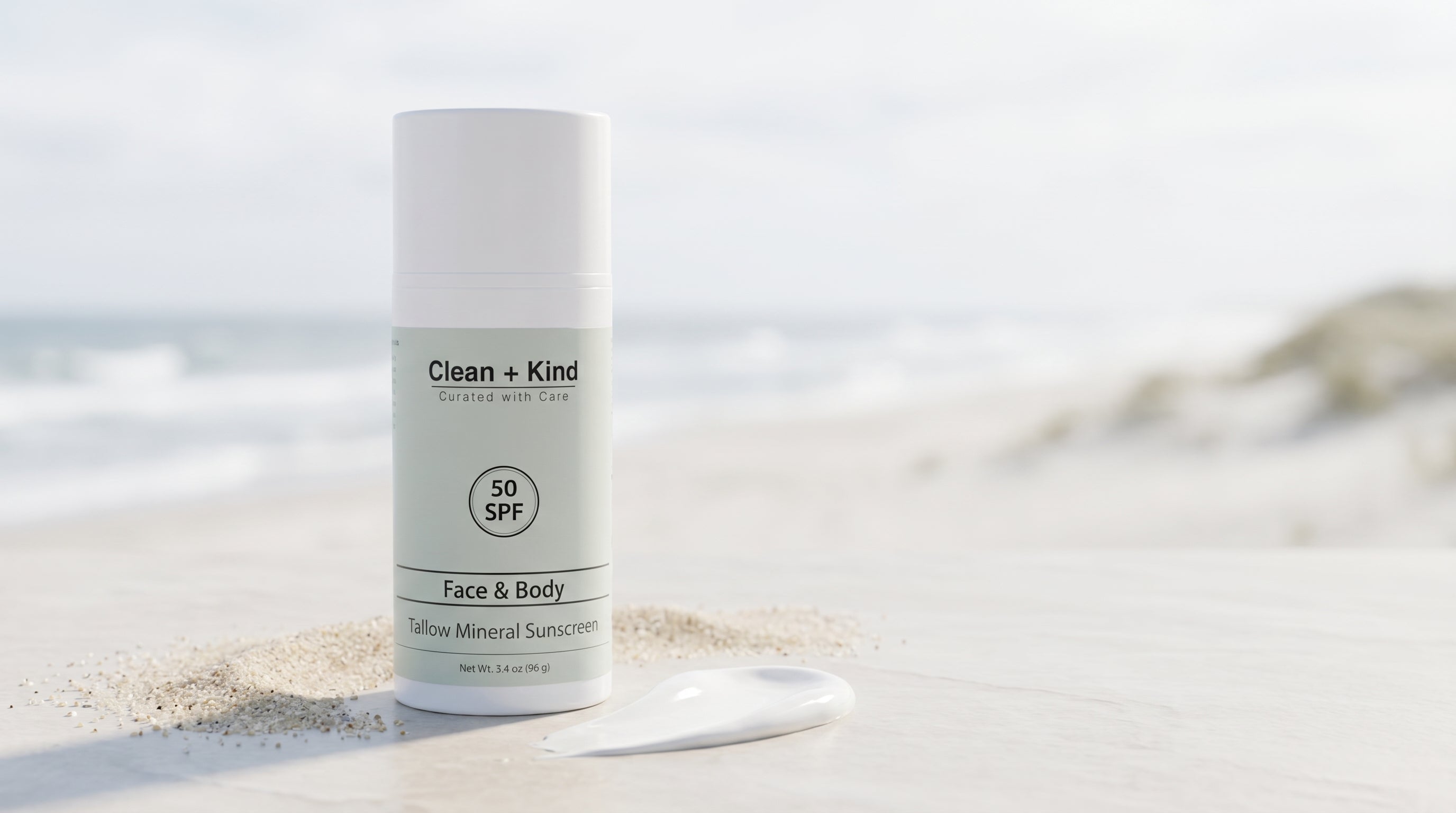 Clean + Kind Tallow Mineral Sunscreen SPF 50 bottle on a sunny beach beside a swatch of mineral cream