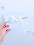 A white cotton mini pinwheel bow attached to a tan headband, displayed on a branded Evie Pearl Handmade backing card.