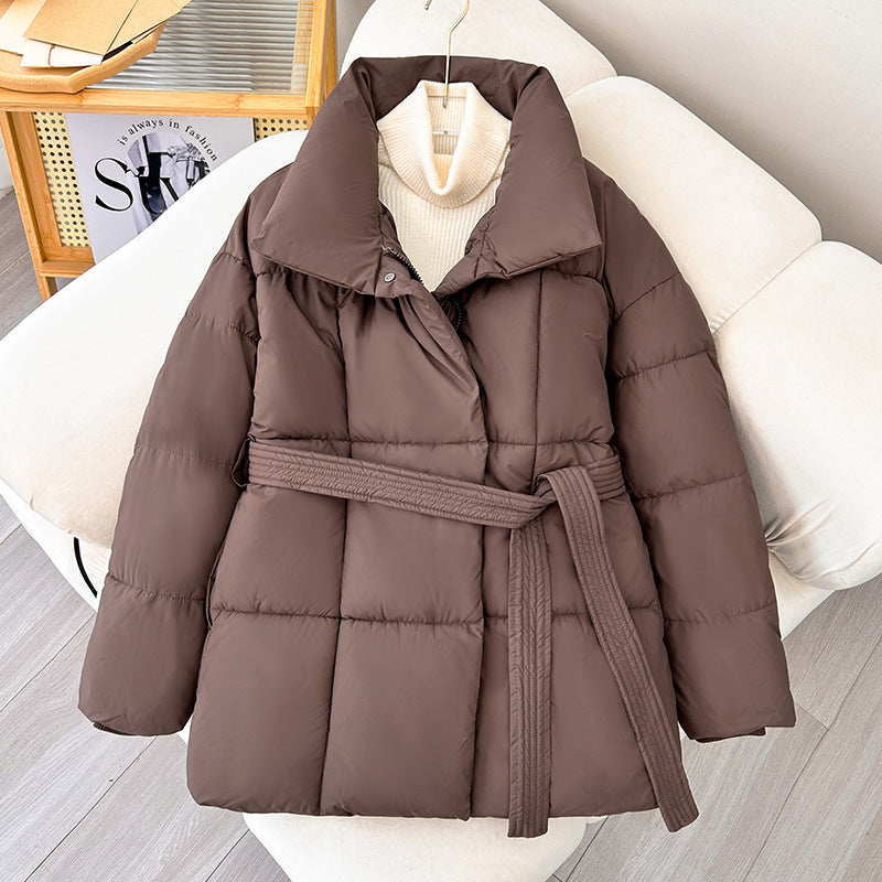 Brown women's puffer parka with high stand collar and belted waist, on a white couch.