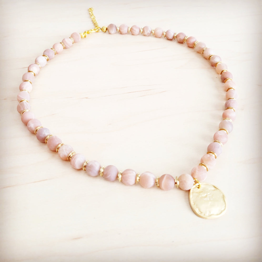 Frosted sunstone beaded collar necklace with a central matte gold-tone pendant on a light wood background