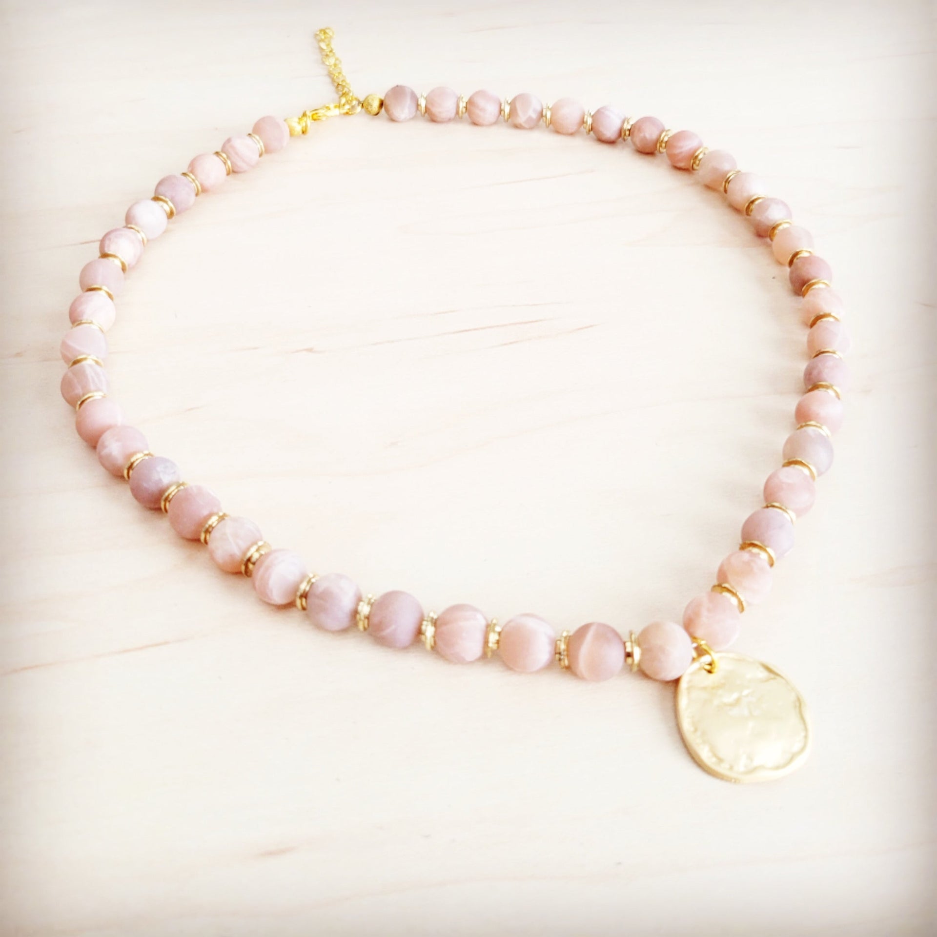Frosted sunstone beaded collar necklace with a central matte gold-tone pendant on a light wood background