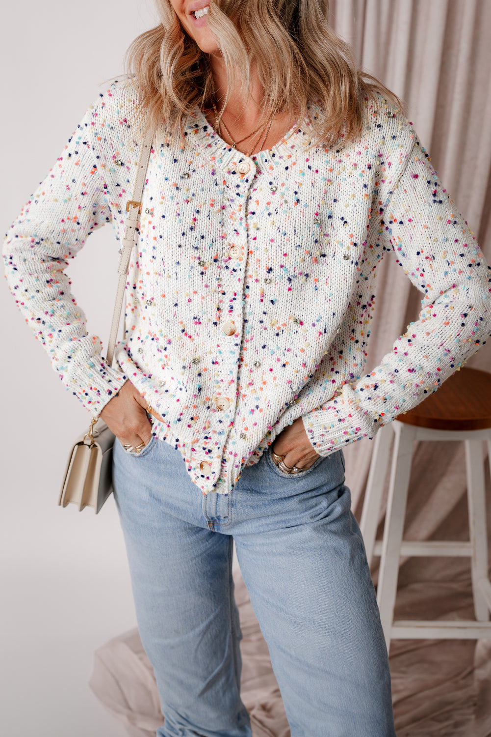 A woman wearing a white button-front cardigan sweater with a multicolor confetti pattern and rhinestone embellishments, styled with light wash jeans.