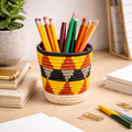 Handwoven Ugandan pencil basket with geometric patterns in yellow, red, black, and cream on a wooden desk.