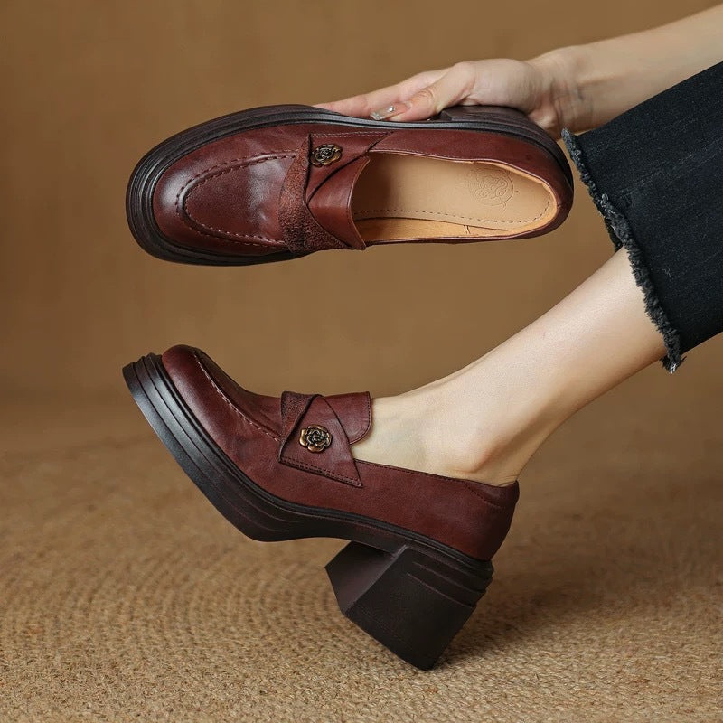 A pair of maroon leather chunky heel loafers featuring a vintage-inspired decorative emblem, shown held in hand and worn on foot.