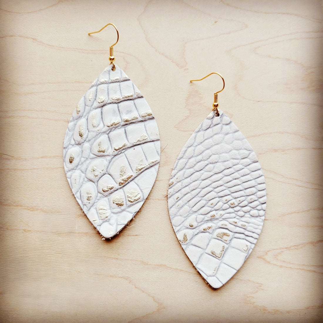 A pair of white leather oval earrings with a gold-tone gator print pattern and gold-tone fish hooks resting on a wood background.