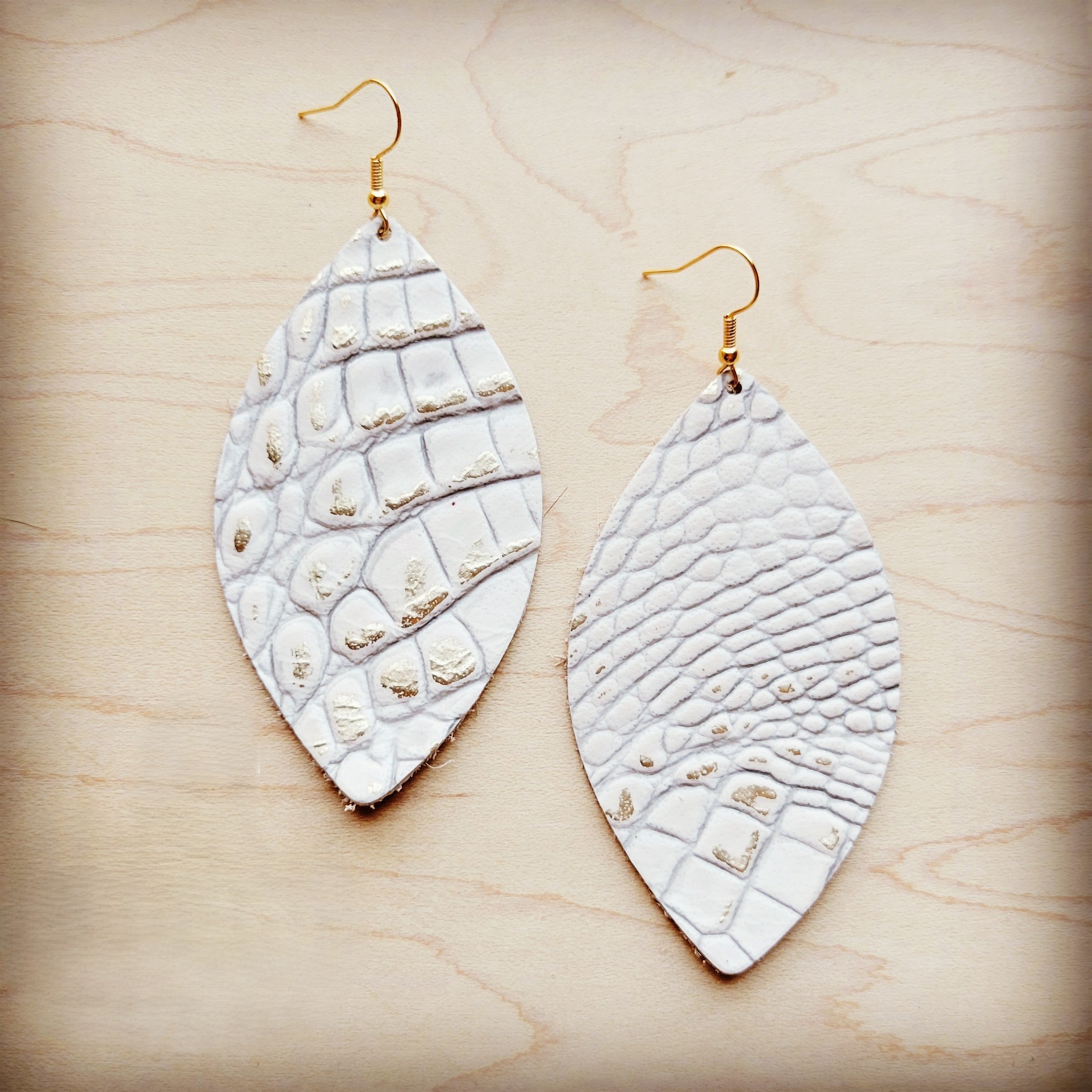A pair of white leather oval earrings with a gold-tone gator print pattern and gold-tone fish hooks resting on a wood background.