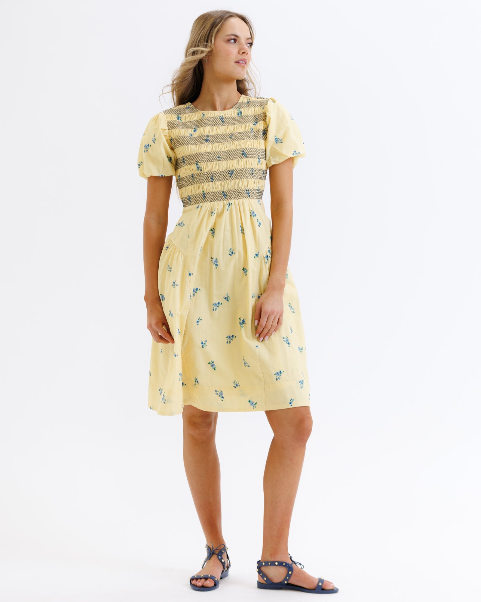 Model wearing a yellow cotton midi dress with blue floral print, smocked bodice, and short puff sleeves.