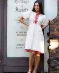 Model wearing a knee-length cream cotton dress with short puff sleeves and red tie details on the bodice.