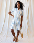 Woman wearing a blue and white vertical striped cotton shirt dress with a collared neckline and pleated skirt.