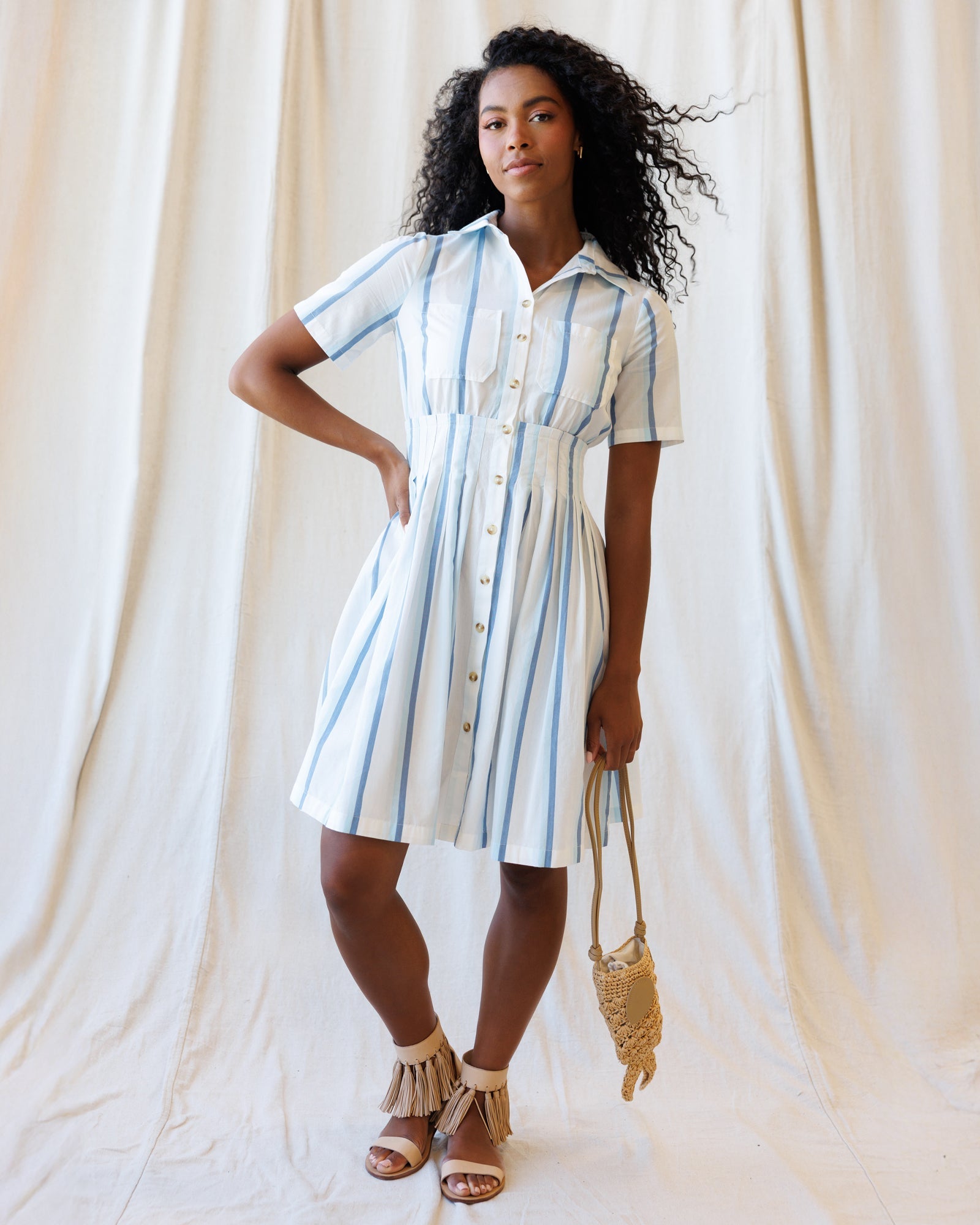 Woman wearing a blue and white vertical striped cotton shirt dress with a collared neckline and pleated skirt.