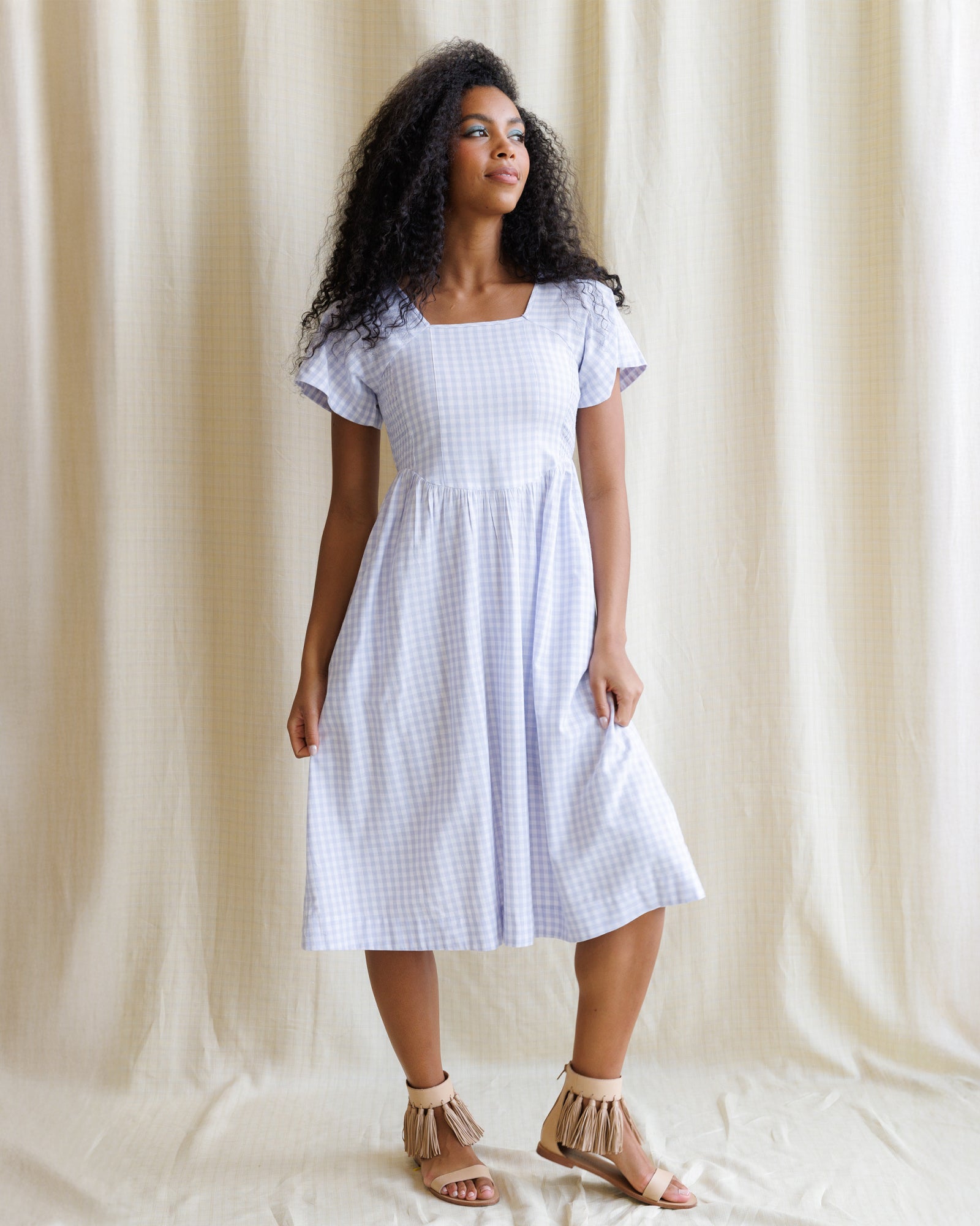 A woman wearing a lavender and white gingham midi dress with flutter sleeves and a square neckline in a garden setting.
