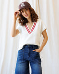 A woman wearing a white preppy knit vest with red varsity stripe trim on a V-neckline