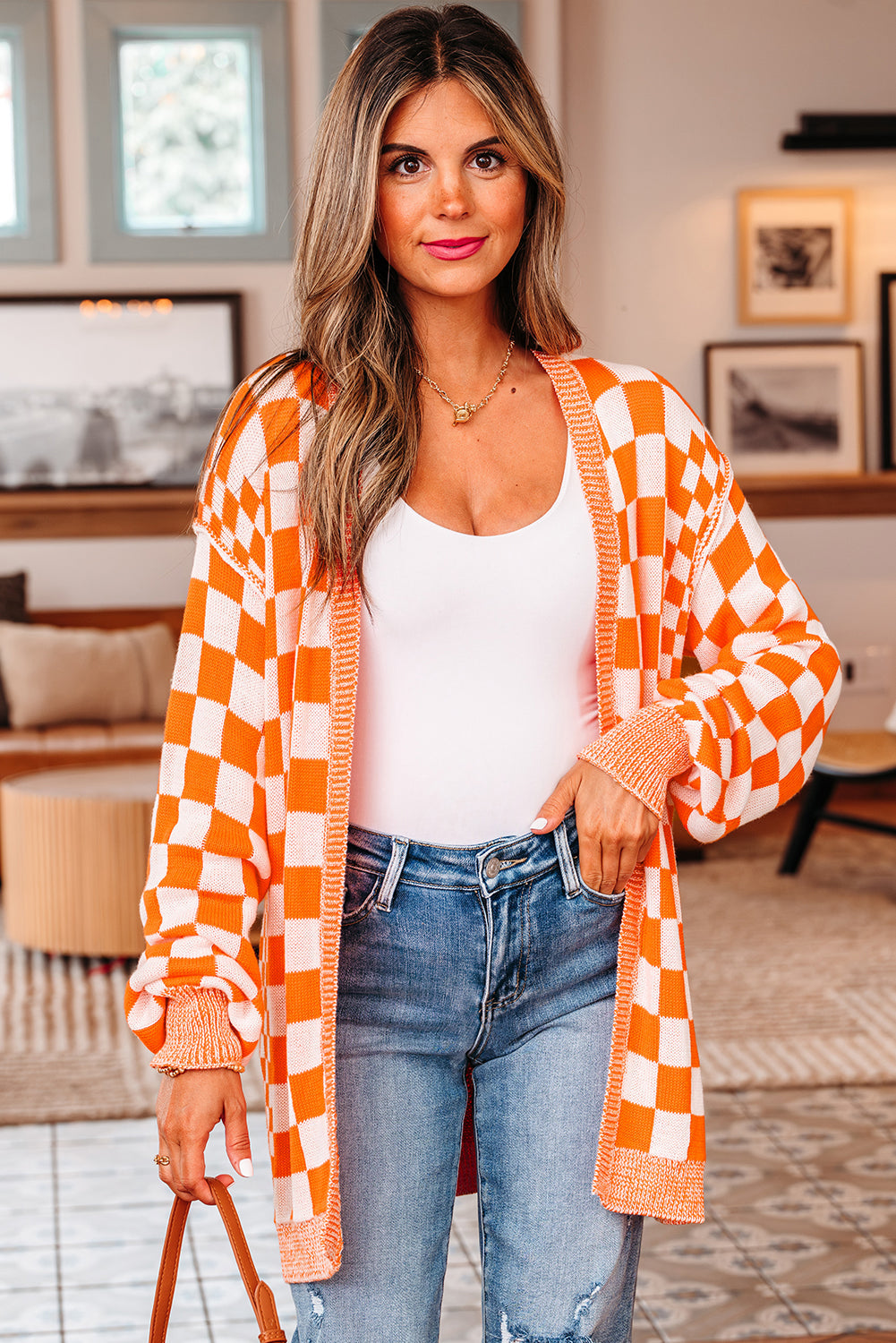 A woman wearing an orange and white checkered open-front knit cardigan with drop shoulders over a white tank top and denim jeans.