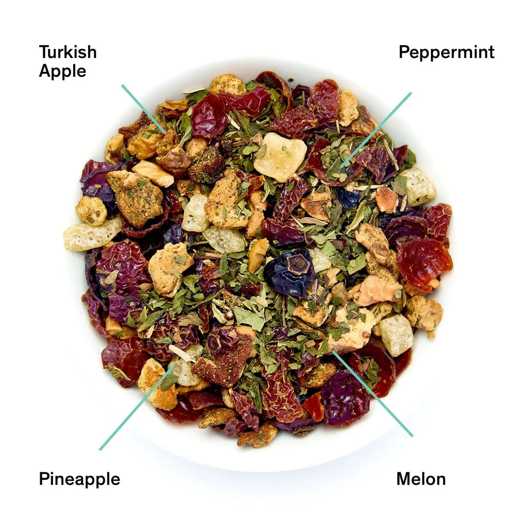A black package of August Uncommon Tea Capri blend next to a small white bowl filled with the loose leaf tea, showing dried melon, peppermint, apple, and pineapple pieces.