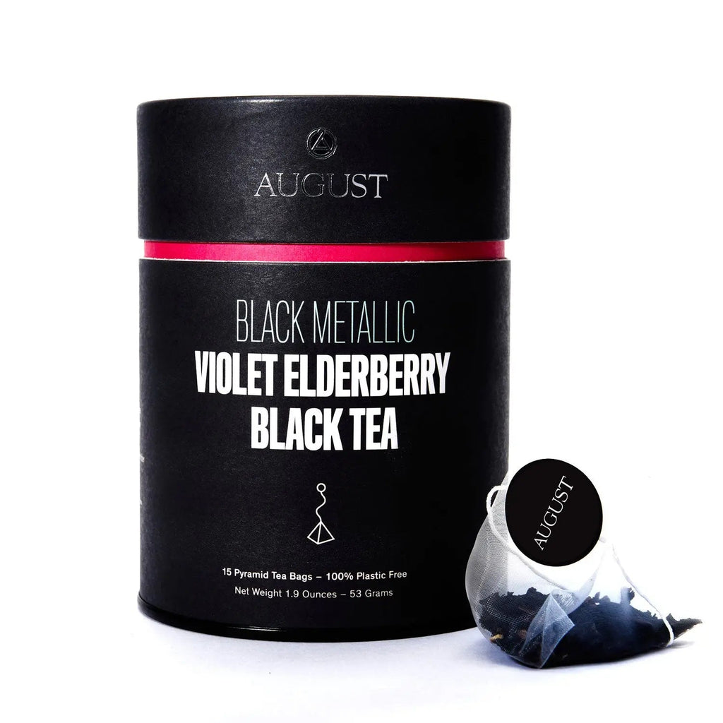 A black cylindrical container of Black Metallic tea next to a single pyramid tea bag, alongside a bowl of the loose leaf tea blend showing dark tea leaves, blue cornflowers, and fruit bits.