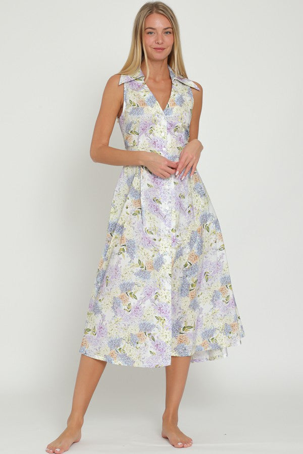 A woman wearing a white and light blue floral sleeveless midi dress with a collar, button-down front, and tie-back detail.