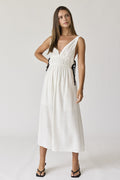 A woman wearing a white V-neck maxi dress with black side ties, standing against a neutral studio background.
