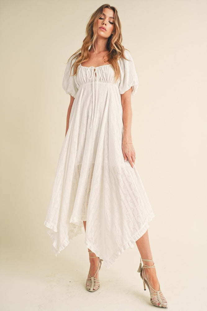 A model standing wearing a white flowy midi dress with puff sleeves and a tie-front detail against a neutral studio background.