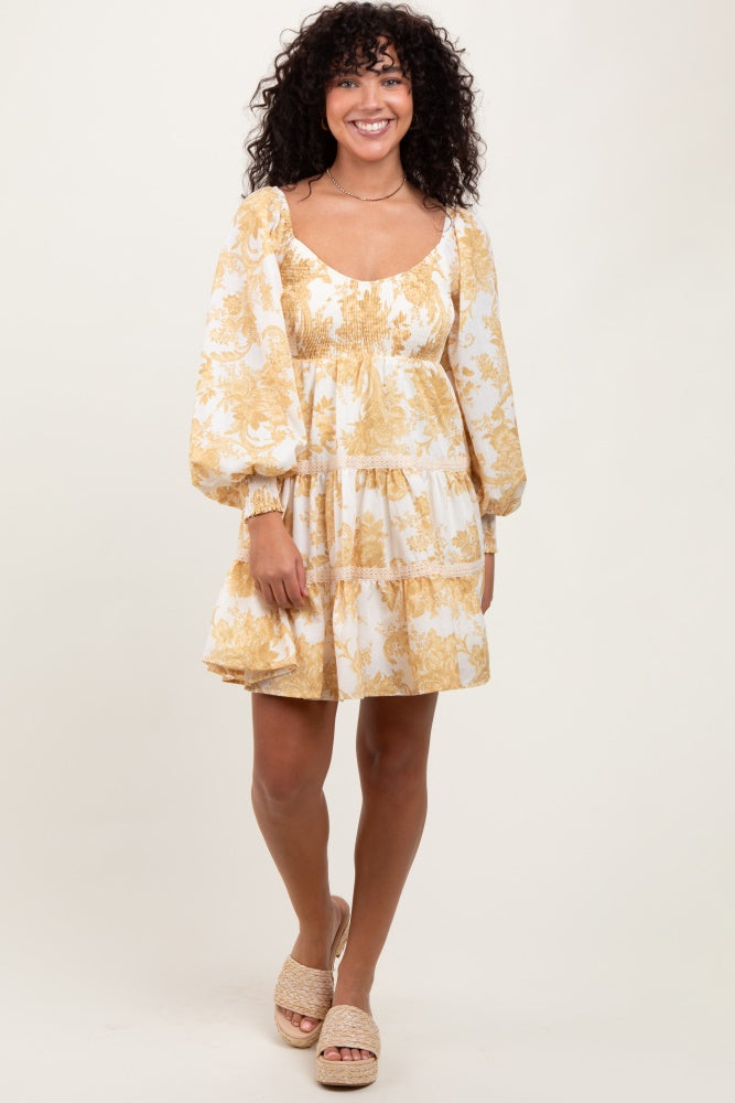 A woman wearing a yellow and white floral print mini dress with smocked bodice, square neckline, bubble sleeves, and lace trim detailing on the tiered skirt.