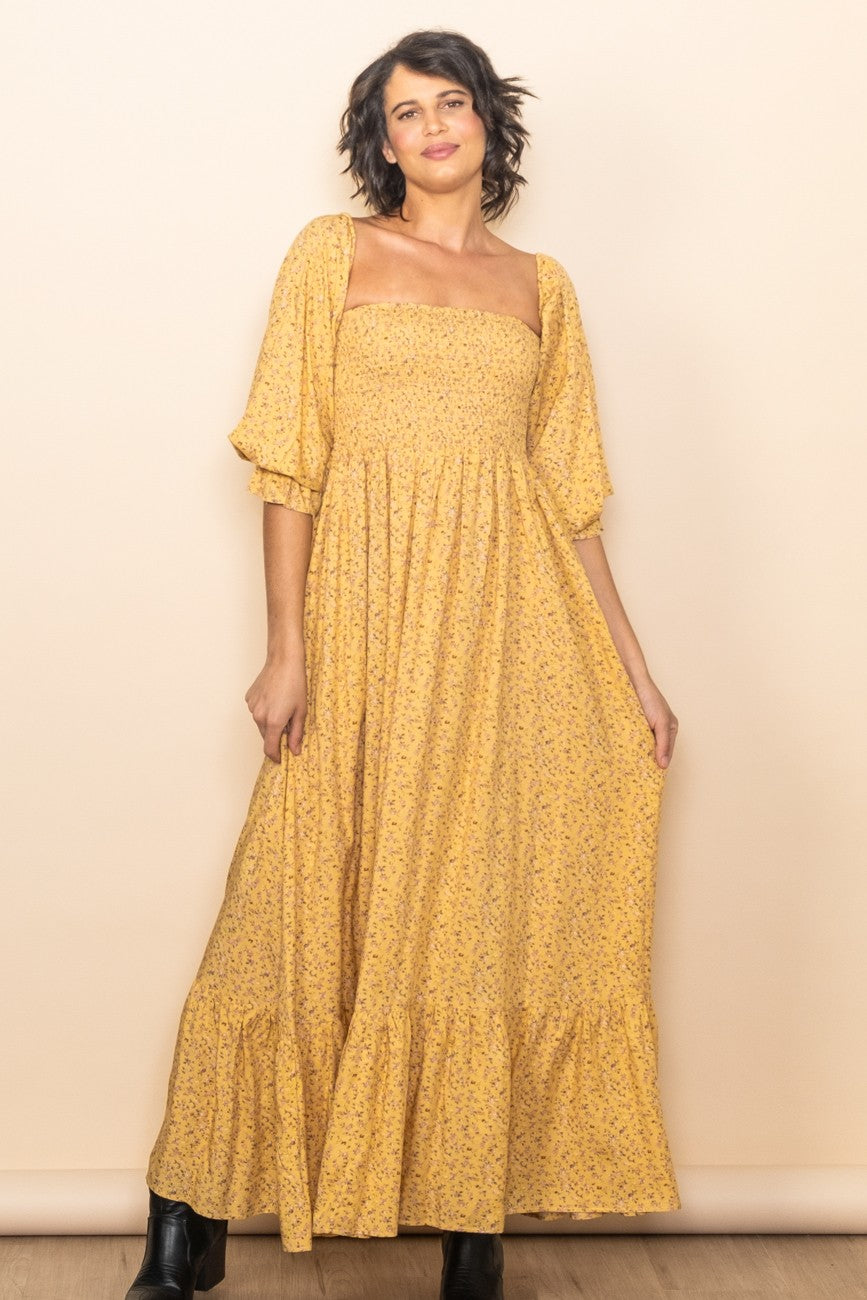 A woman wearing a yellow floral maxi dress with a smocked bodice and puff sleeves, standing against a neutral studio backdrop.