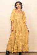 A woman wearing a yellow floral maxi dress with a smocked bodice and puff sleeves, standing against a neutral studio backdrop.