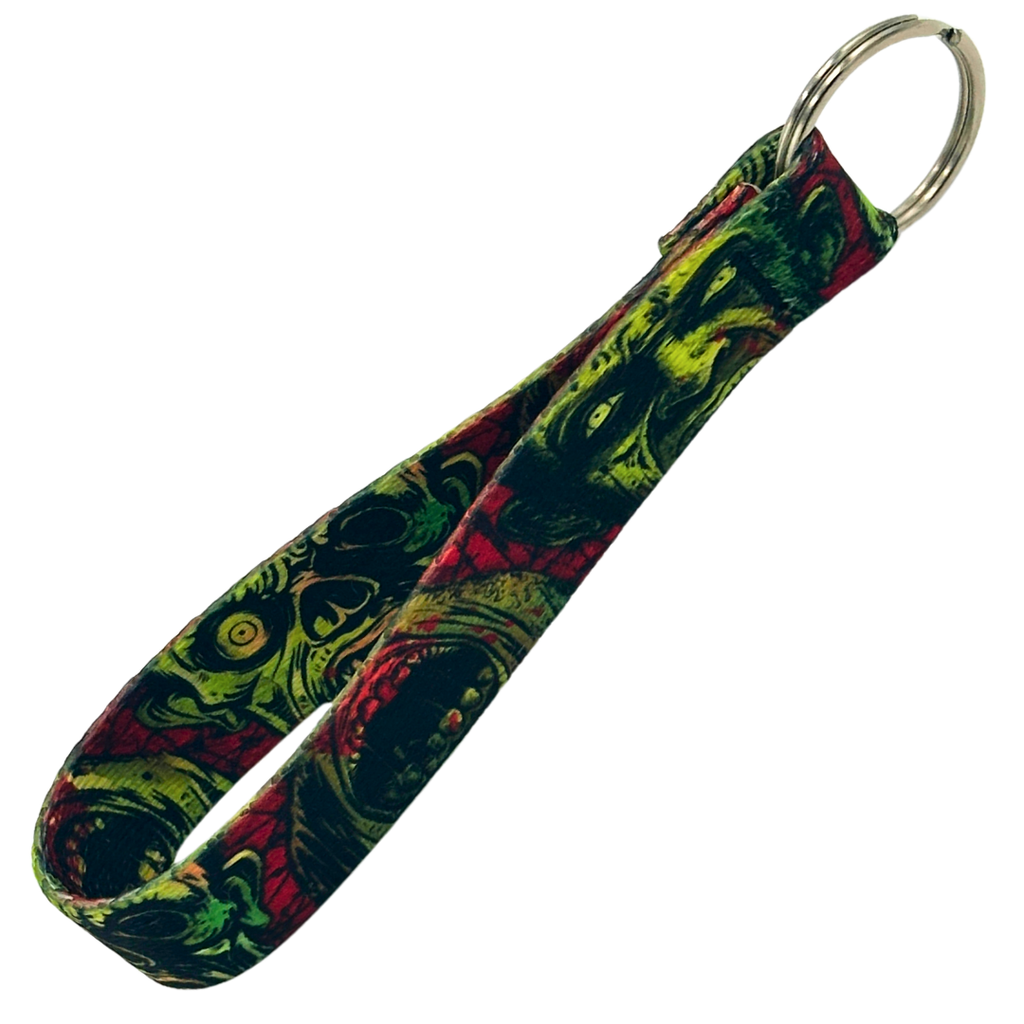 A fabric wristlet key fob featuring a green and red zombie graphic print with a metal key ring attachment against a graveyard background.