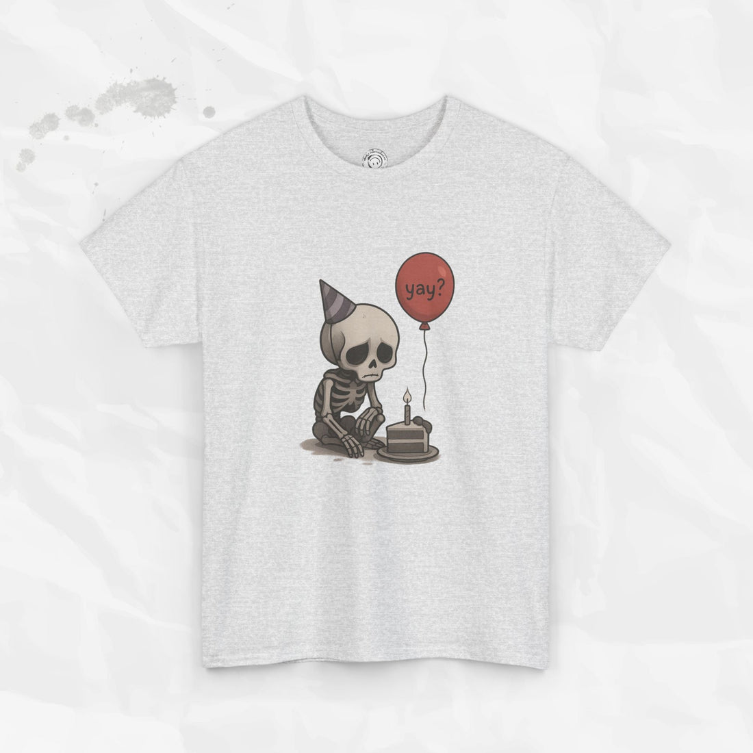 A close-up shot of a t-shirt featuring a sad skeleton wearing a party hat with a balloon and a birthday cake, available in heather grey or coral red.