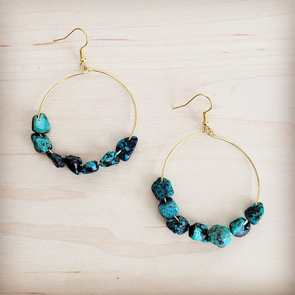 A pair of gold-tone hoop earrings featuring raw turquoise beads on a light wooden background