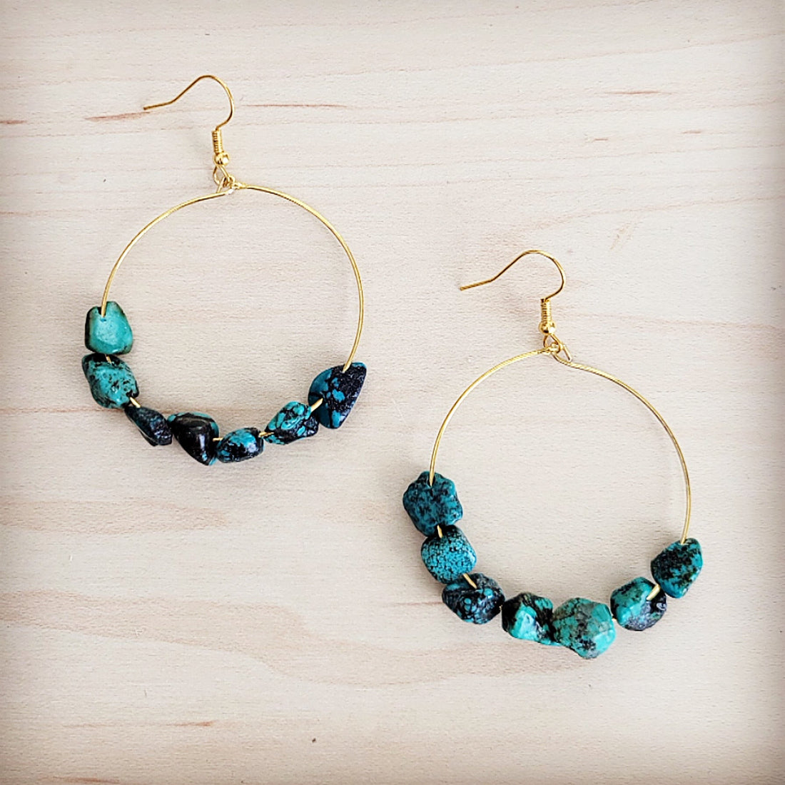 A pair of gold-tone hoop earrings featuring raw turquoise beads on a light wooden background