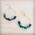 A pair of gold-tone hoop earrings featuring raw turquoise beads on a light wooden background