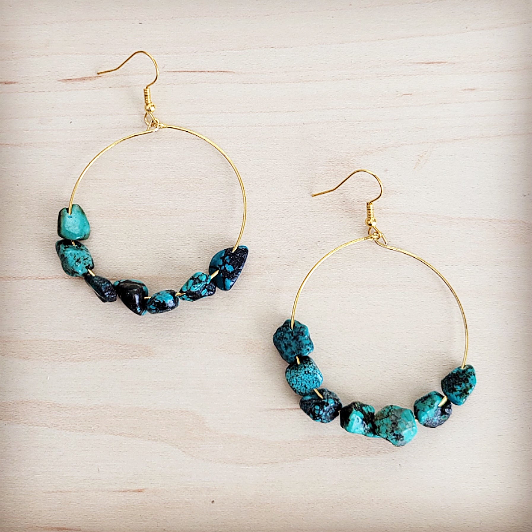 A pair of gold-tone hoop earrings featuring raw turquoise beads on a light wooden background