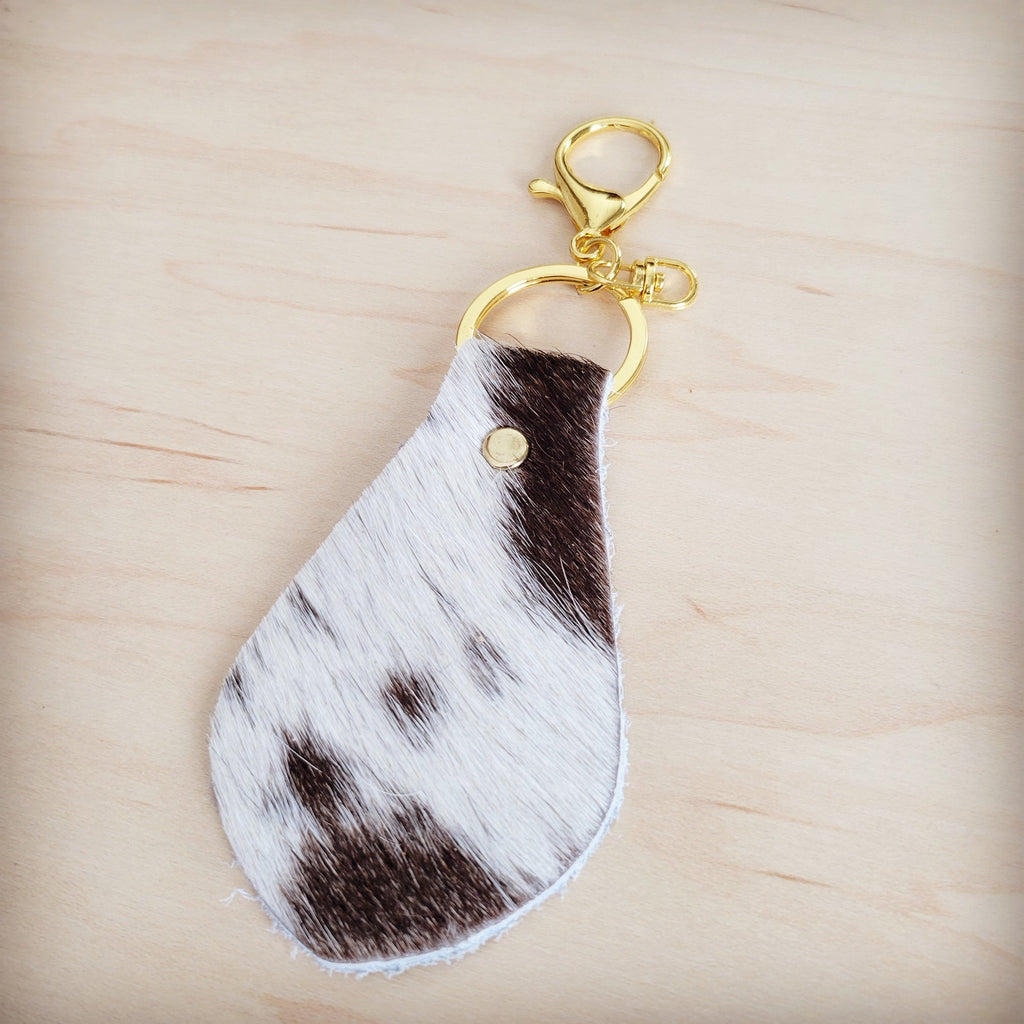 A teardrop-shaped brindle cowhide leather keychain featuring hair-on-hide texture and a gold-tone clasp on a light wooden background.