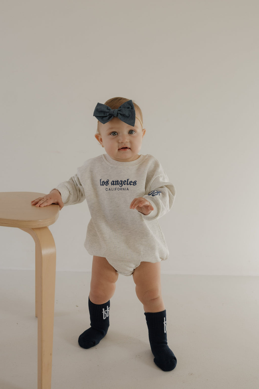 Baby wearing a white long-sleeve outfit with 'Los Angeles California' text, standing next to a wooden table.
