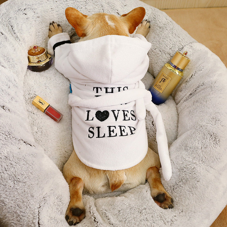 A French Bulldog wearing a white hooded pet bath robe with the text This Loves Sleep on the back while lying in a plush bed.