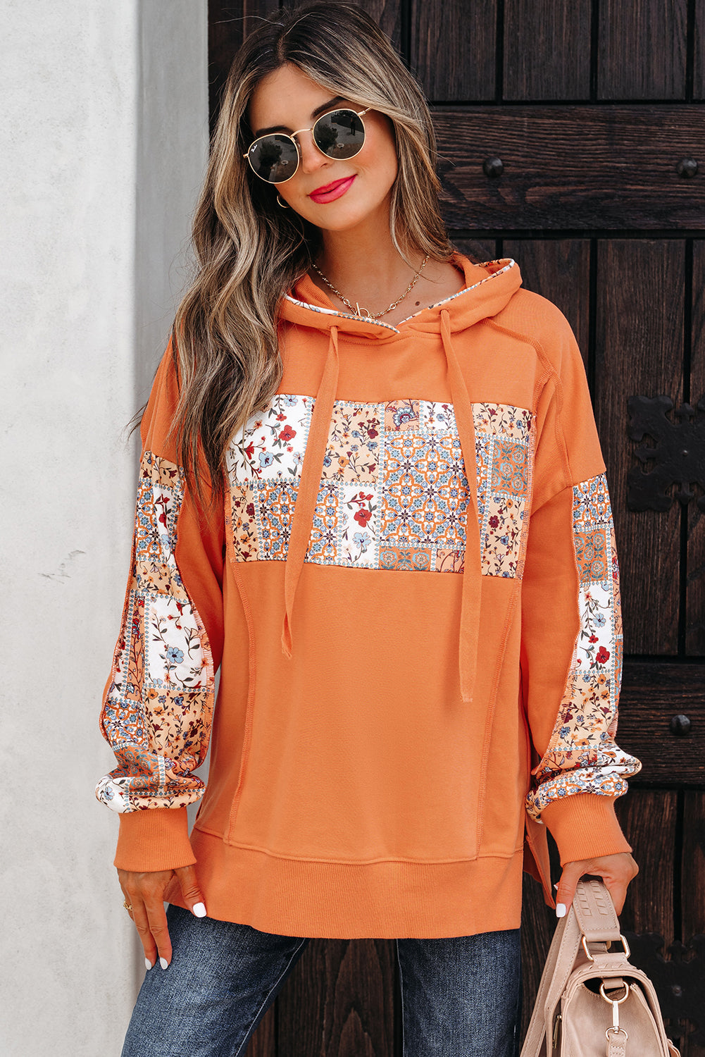 Woman wearing a vibrant pumpkin orange floral patchwork hoodie with side slits and a drawstring hood.