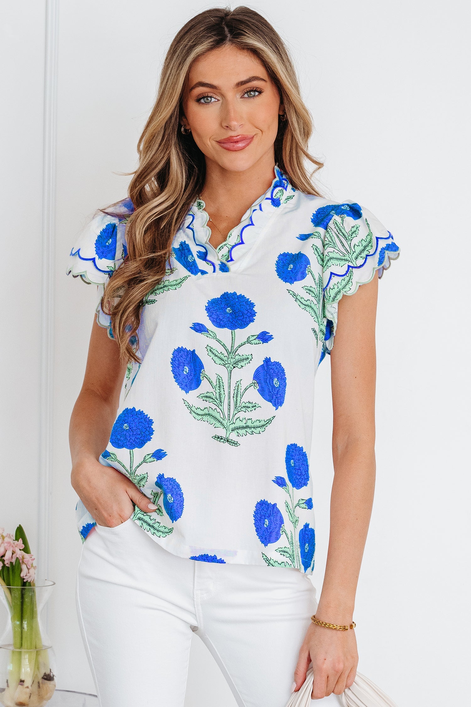 Model wearing a white blouse with a blue and green wild flower print, featuring a scalloped neckline and short sleeves with blue contrast trim.