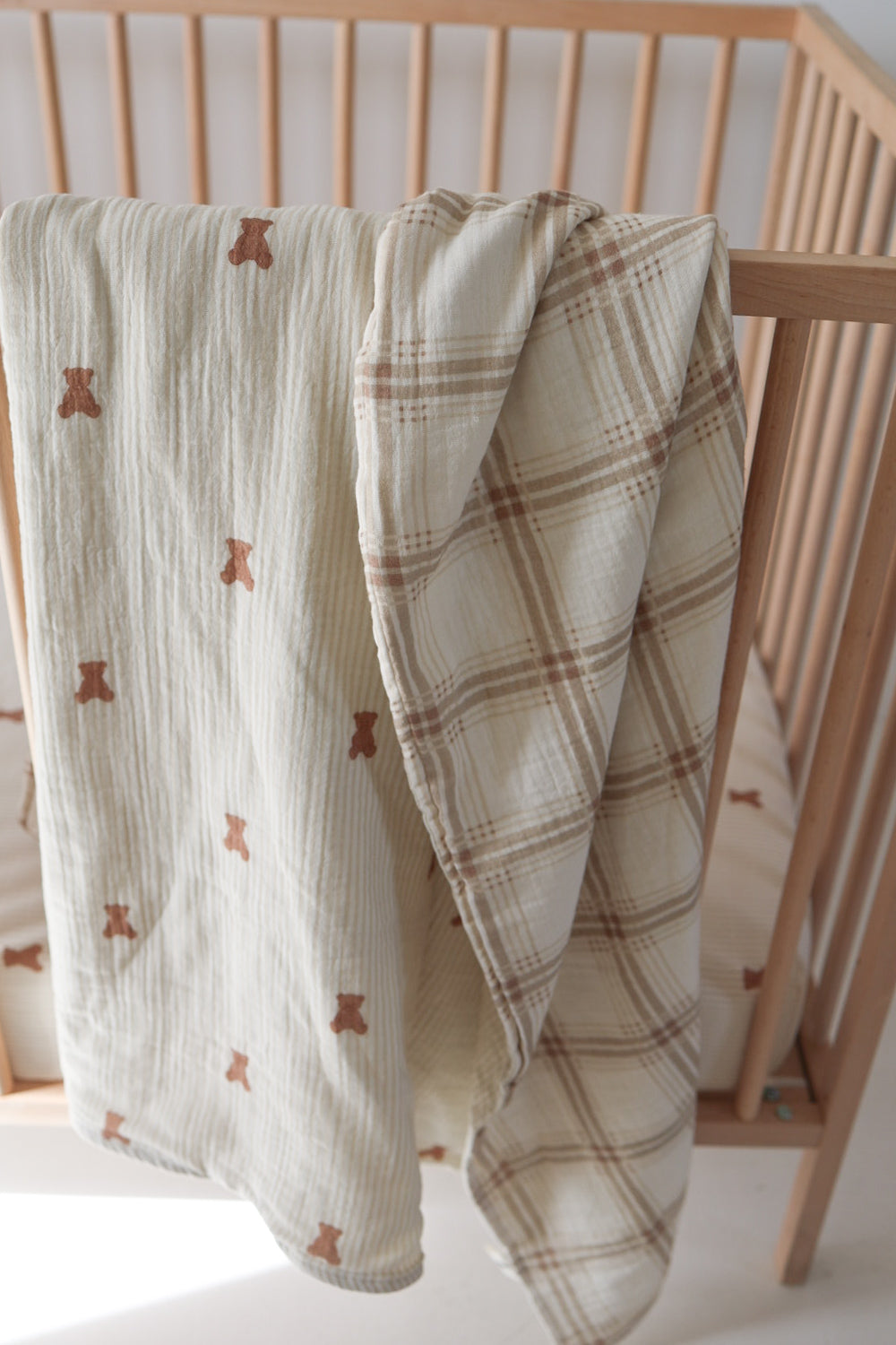 Reversible baby muslin quilt folded over a wooden crib, showing a teddy bear print on one side and a beige plaid print on the other.