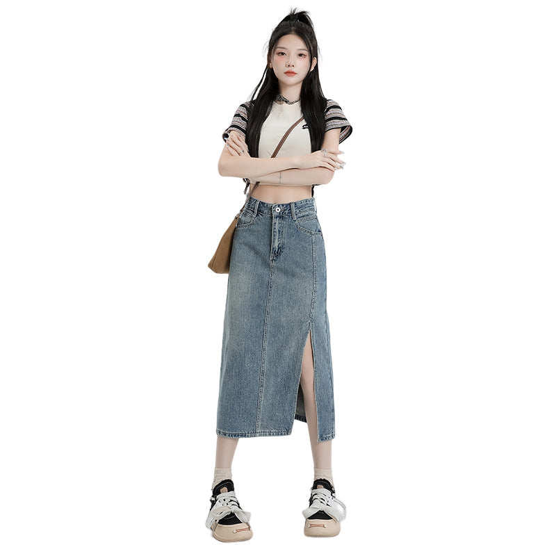 A woman wearing a blue high-waisted A-line denim midi skirt with a front slit, paired with a white crop top and sneakers, posed against a white background.