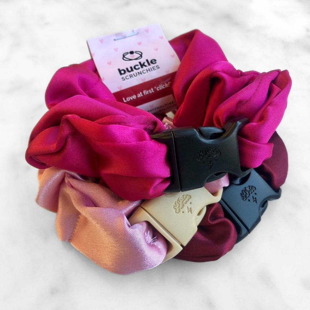 A set of three satin buckle scrunchies in fuchsia, berry, and pale-pink colors for Valentine's Day.