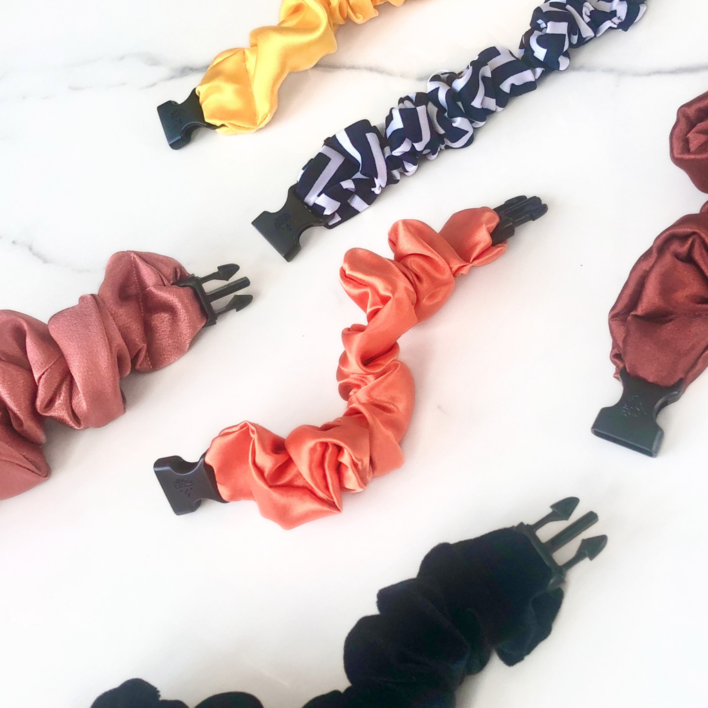 A selection of minimalist satin buckle scrunchies in black, sienna, orange, and yellow.