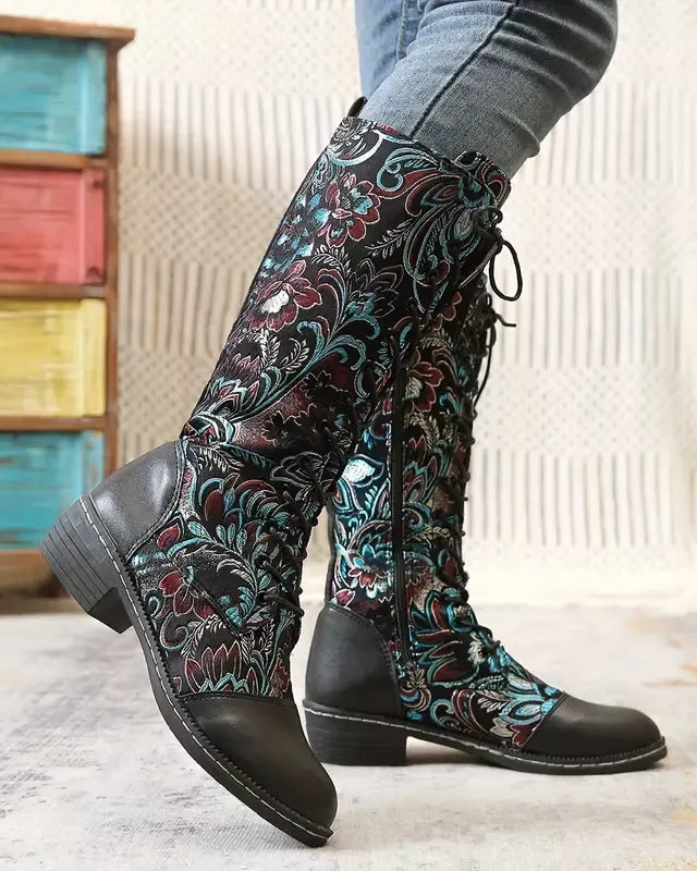 Black knee-high boots with floral pattern worn by a person, with a colorful wooden drawer set in the background.