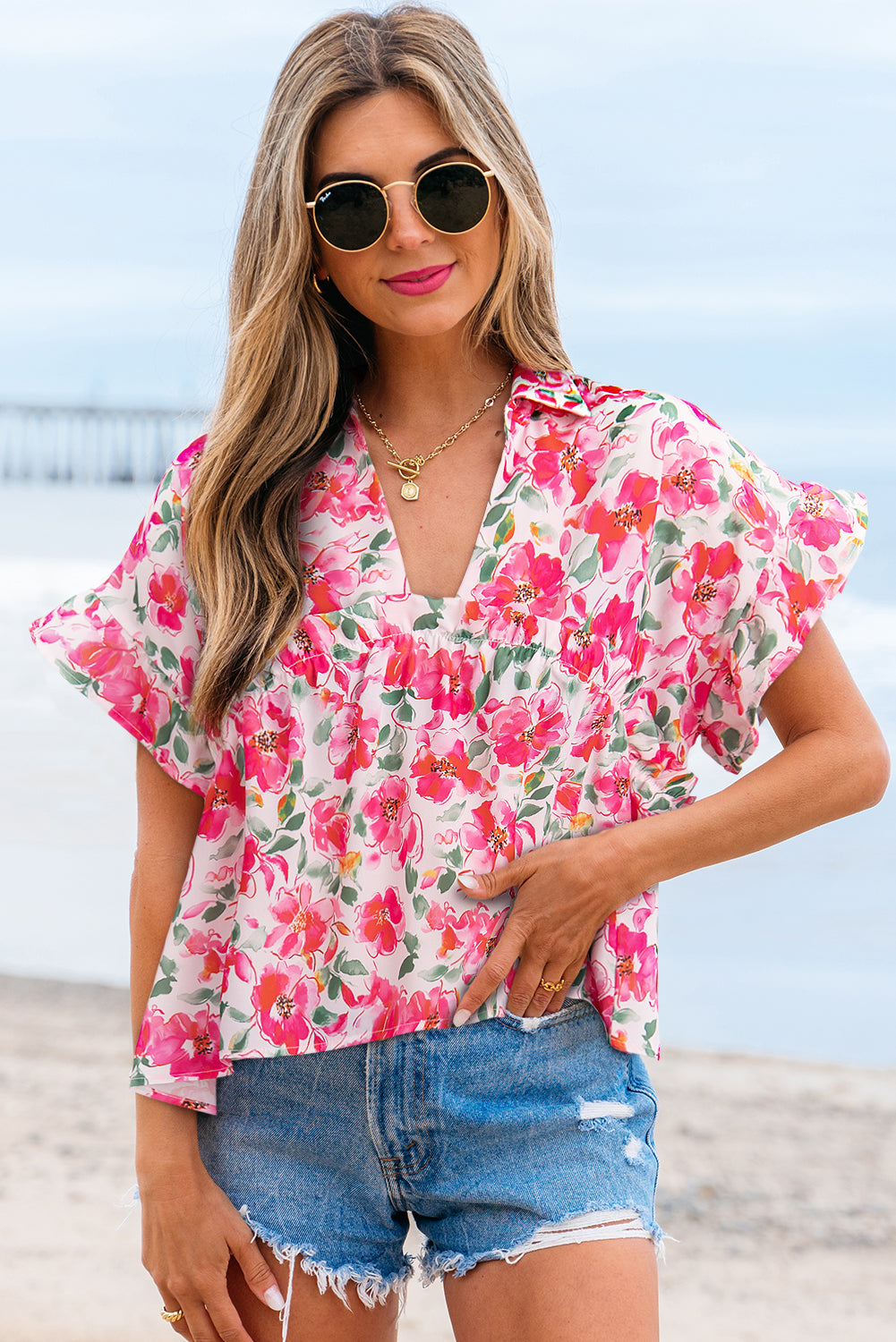 Model wearing a pink and white floral patterned short sleeve blouse with ruffled sleeves and a v-neckline on a sandy beach.