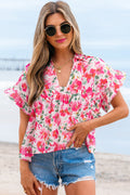 Model wearing a pink and white floral patterned short sleeve blouse with ruffled sleeves and a v-neckline on a sandy beach.