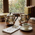 A hand-hammered fine silver teapot and matching creamer with sleek black handles and warm metallic base accents.