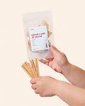 A person holding a transparent bag filled with various sizes of natural wood wicks and metal clips for candle making.