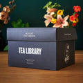 A black Tea Library gift box filled with 30 pouches of gourmet loose leaf tea on a wooden surface with a clear tea scoop.