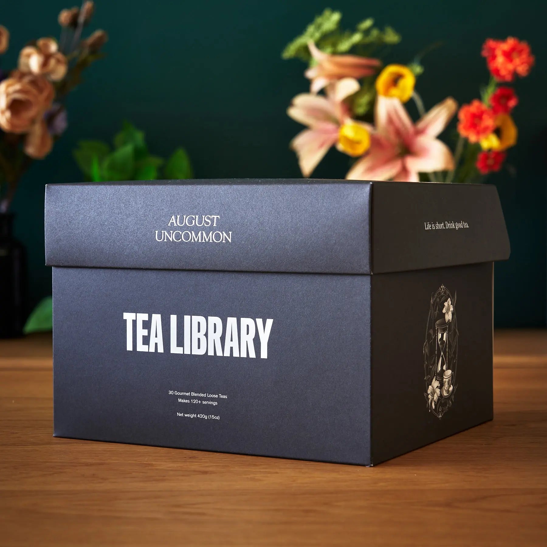 A black Tea Library gift box filled with 30 pouches of gourmet loose leaf tea on a wooden surface with a clear tea scoop.
