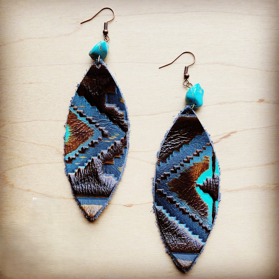 A pair of narrow oval-shaped genuine leather earrings featuring a blue and brown Navajo pattern with a turquoise stone accent on brass hooks.