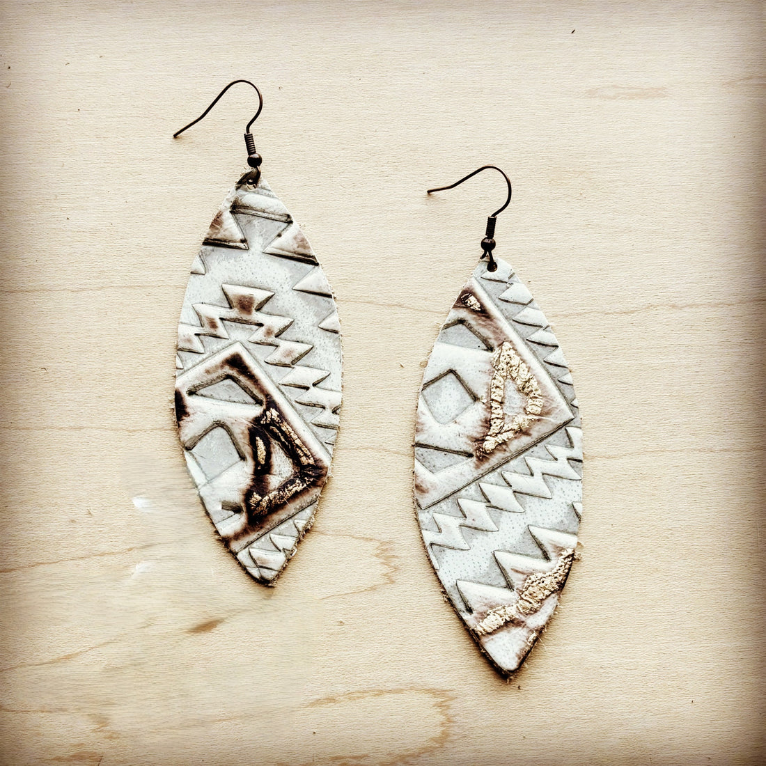 A pair of narrow oval leather earrings featuring a white and brown Aztec geometric pattern with gold leaf accents on a wooden background.