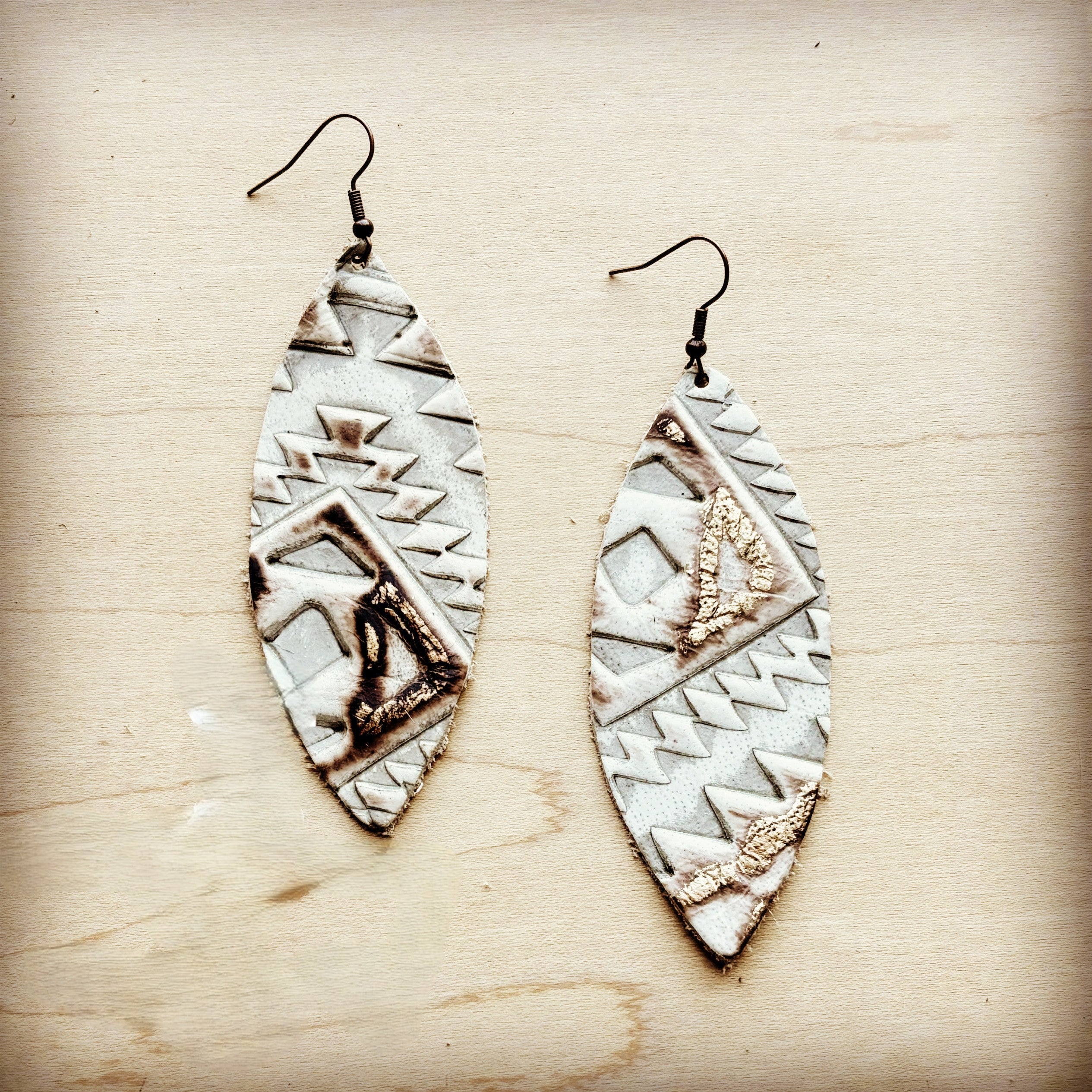 A pair of narrow oval leather earrings featuring a white and brown Aztec geometric pattern with gold leaf accents on a wooden background.